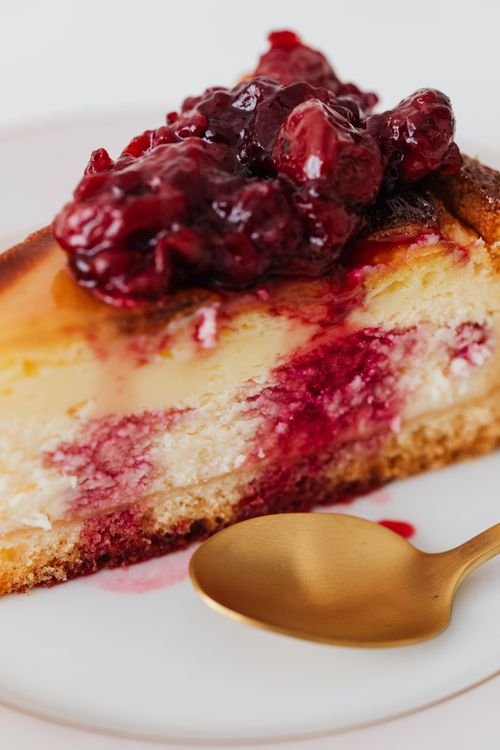 Slice of cherry cheesecake with glossy berry topping and a gold spoon on a white plate